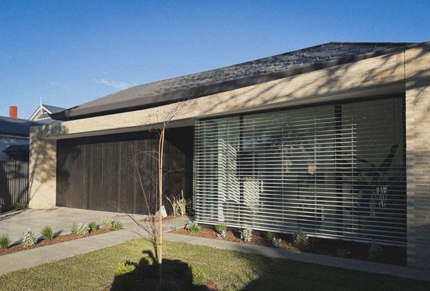 Single-storey contemporary residential facade viewed from the street, with pale textured brick walls, a dark low-pitched tiled roof, a broad dark garage door, and large horizontal-louvred glazing. A simple landscaped front garden and concrete path occupy the foreground.