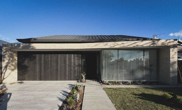 Single-storey contemporary residential facade with a low-pitched dark metal roof, pale textured masonry walls, a large dark timber garage door, and broad horizontal glazing fitted with external venetian blinds. A concrete driveway and path lead through a narrow landscaped garden bed to the recessed entry. The frontage has a clean, restrained modern composition with strong horizontal lines.