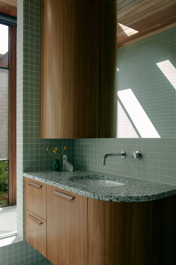 A close-up view of a contemporary bathroom vanity with a curved green-speckled stone benchtop, integrated white basin, timber cabinetry, and a wall-mounted chrome tap. Small square pale green tiles cover the walls and floor, while a large timber-framed mirror and timber-lined ceiling add warmth. Natural light enters through a tall window and creates strong geometric shadows on the tiled wall.