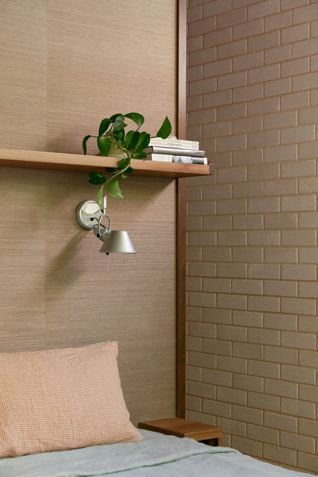 Close-up of a warm, compact bedroom corner featuring a bed with a pale blue-grey cover and peach check cushion, a timber floating shelf with books and a trailing plant, a small articulated wall lamp, and contrasting textured timber wall panelling beside exposed brown brickwork.