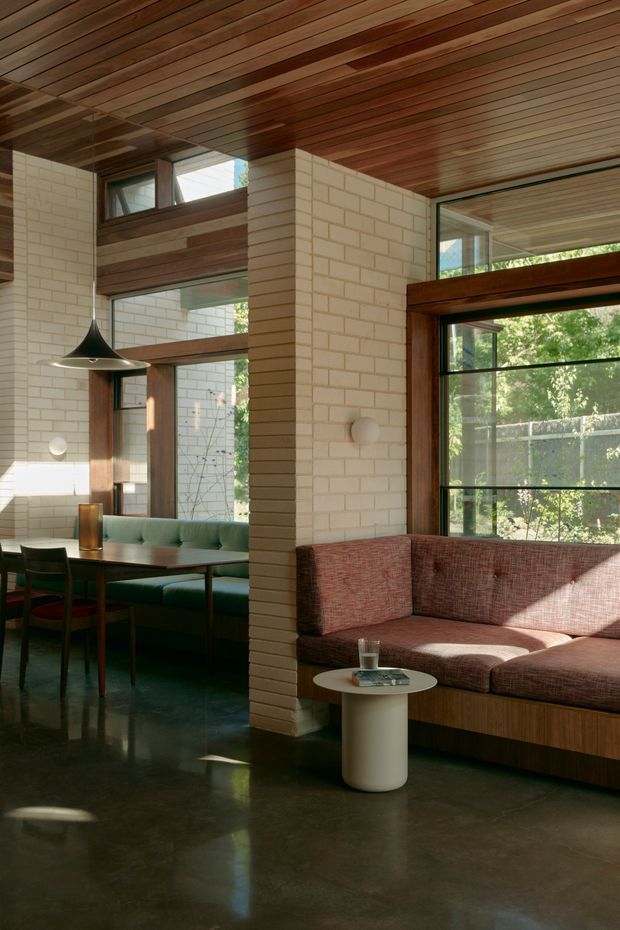 Interior view of a warm, mid-century-inspired open-plan living and dining area. The space features exposed dark timber slat ceilings, cream painted brick walls, dark polished concrete flooring, extensive timber-framed glazing, a built-in rust-coloured sectional sofa, and a green upholstered dining banquette with a timber dining table. A sculptural black pendant light and small round side table complete the refined, natural-toned interior.