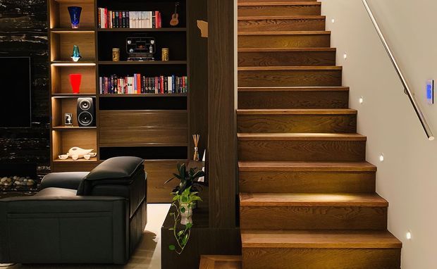 A moody contemporary interior showing a dark timber built-in media and bookshelf wall beside a warm timber staircase. The shelving displays books, audio equipment and decorative objects, while a black sectional sofa occupies the foreground. Recessed shelf lighting and small stair lights add warmth, with a slim metal handrail on the pale wall.