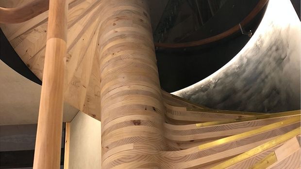 Close-up interior architectural detail featuring a sculptural, flowing timber staircase or balustrade with layered curved wood grain, a rounded timber support column, and dark overhead structural elements. The organic joinery creates a dramatic, highly crafted focal point.