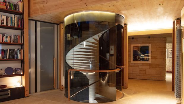 Interior view of a warm, timber-lined contemporary home featuring a striking sculptural spiral staircase enclosed in a cylindrical glass shaft. Built-in bookshelves occupy the left wall, while a stone-clad wall and framed artwork are visible beyond the stair enclosure. Recessed and concealed lighting create an atmospheric, refined ambience.