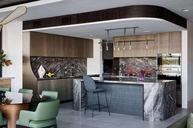 A sophisticated contemporary kitchen and bar area with dark timber cabinetry, dramatic veined stone splashbacks and island cladding, integrated appliances, and a large island with textured dark panel detailing. Sculptural pendant lighting hangs above the island, while green upholstered dining chairs and a small round timber table are visible in the foreground. The space has a refined, high-contrast palette and a curved ceiling bulkhead with dark ventilation grilles.