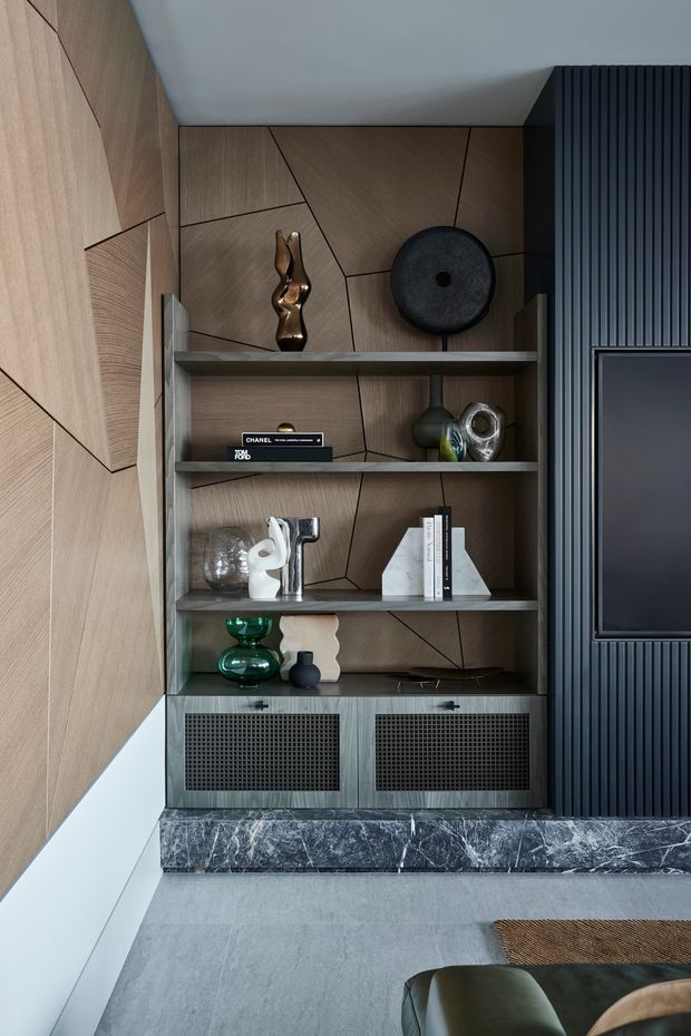 A refined contemporary living room media wall featuring a built-in open shelving unit with dark metal framing, decorative objects, books, and integrated speaker cabinets. The wall combines geometric timber veneer panelling with black vertical fluted panelling and a wall-mounted television. A dark marble plinth runs beneath the joinery, with grey tiled flooring and partial seating visible in the foreground.