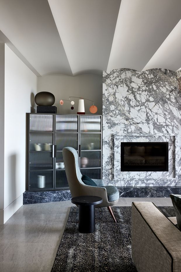 A refined contemporary living room featuring a dramatic grey-and-white marble fireplace wall with a recessed black fireplace, dark ribbed-glass built-in cabinetry, and sculptural lounge furniture. The room has a high sculpted ceiling, pale stone flooring, and a dark textured area rug, creating a luxurious and sophisticated atmosphere.