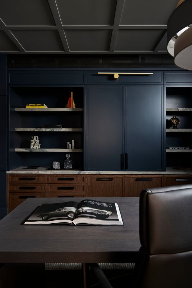 A moody, refined home office featuring dark navy built-in cabinetry with open display shelving, bronze or brass hardware, and a warm timber drawer base. A dark timber desk with an open book occupies the foreground, paired with a leather office chair. The ceiling has a coffered grid detail, and a sculptural pendant light is partially visible at the upper right.
