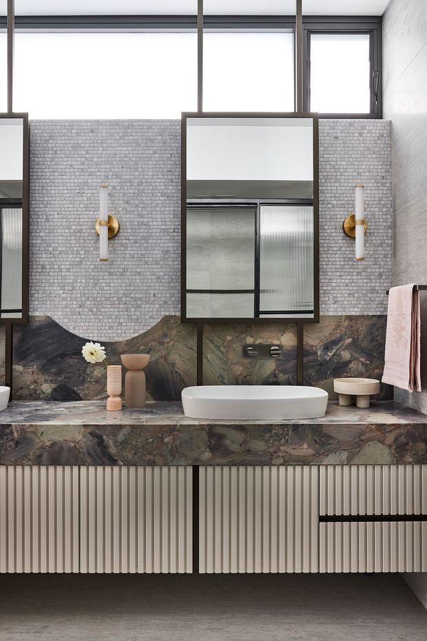 A refined contemporary bathroom featuring a long floating vanity with ribbed light cabinetry, a dark veined stone benchtop and integrated vessel basin. The walls combine small grey brick-pattern tiles with dramatic natural stone panels, framed mirrors, brass-and-white wall sconces, a towel rail with a pale pink towel, and high clerestory windows. The palette is moody and sophisticated with layered grey, brown, cream, brass, and blush tones.