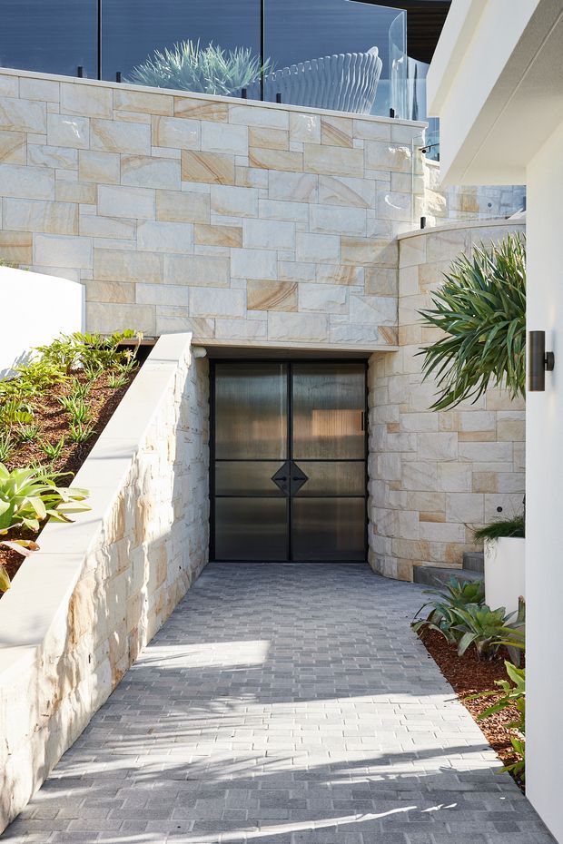 A bright contemporary exterior entry walkway leading to dark metal-framed double doors. Pale stacked natural-stone walls and rendered white architectural elements frame a gently sloping grey paver path, with integrated garden beds and tropical planting. A glass balustrade and upper-level glazing are visible above, creating a refined multi-level residential entrance.