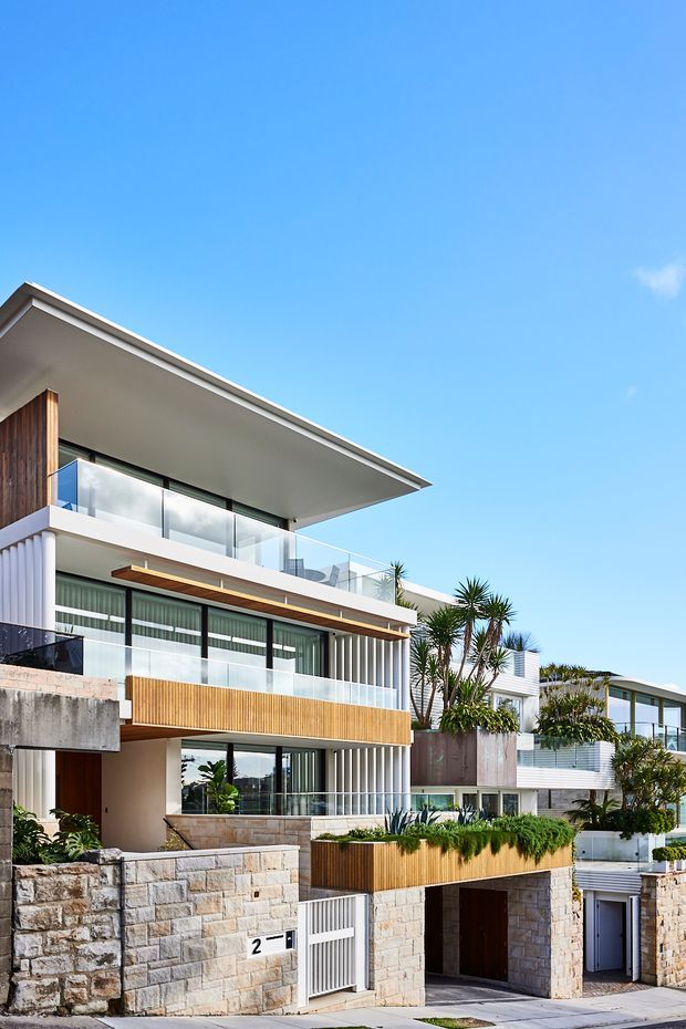 Contemporary multi-storey residential facade on a sloped urban site, featuring projecting flat roof planes, extensive floor-to-ceiling glazing, frameless glass balustrades, warm timber screening and cladding, pale stone masonry retaining walls, and landscaped terraces with sculptural plants. The image is photographed from the street under a clear blue sky.