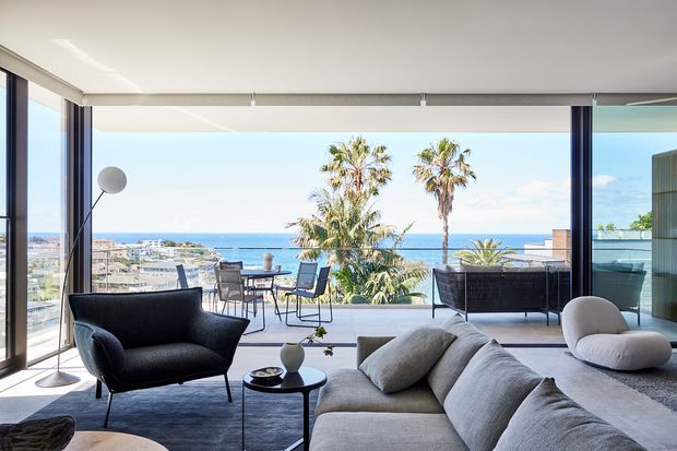 Bright contemporary living room with a large sectional sofa, sculptural lounge chairs, dark side tables, and expansive floor-to-ceiling glazing opening onto a terrace. The view looks over palm trees, a coastal urban setting, and the ocean beyond. The room has a low, refined furniture arrangement and strong indoor-outdoor connection.