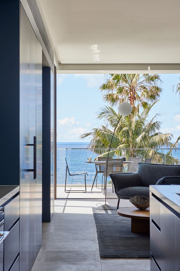 A refined modern living room opening onto a covered balcony or terrace with expansive ocean views. The interior features dark built-in cabinetry, a charcoal upholstered lounge chair, a round timber coffee table, a large dark rug, pale tiled flooring, and full-height glazing. A dining setting and palm trees are visible outside, creating a bright coastal atmosphere.