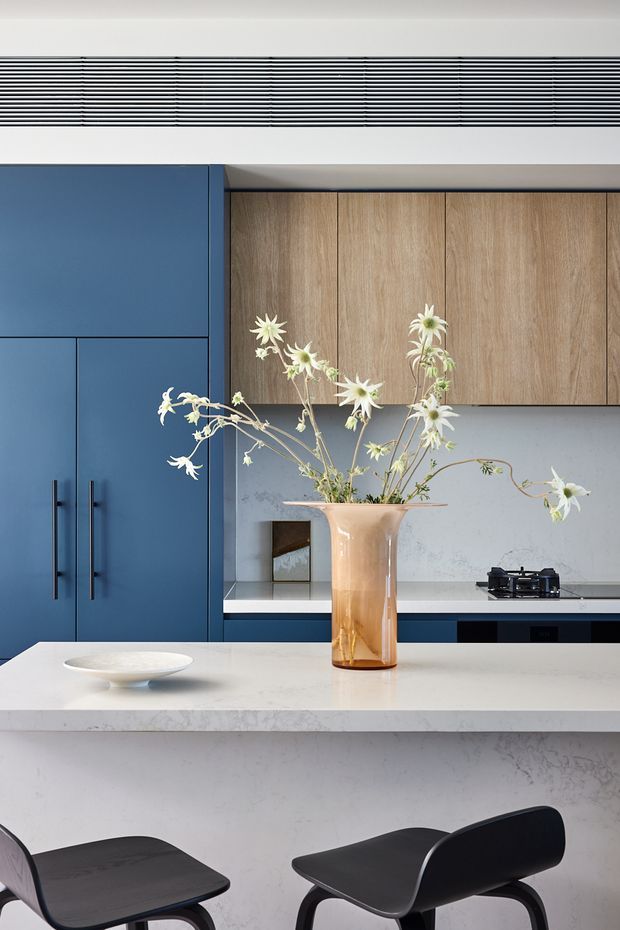 A refined contemporary kitchen featuring deep blue flat-panel cabinetry with black handles, warm wood overhead cupboards, pale stone benchtops and splashback, and a large island with integrated seating. A linear ventilation grille spans the upper wall, while a sculptural vase and white flowers provide a soft decorative focal point. Black bar stools add contrast.