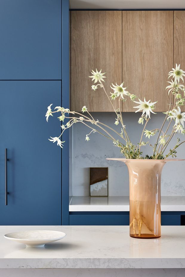 Close-up of a refined kitchen with deep navy-blue cabinetry, tall natural wood overhead cabinets, a pale marble or engineered-stone benchtop and backsplash, and minimalist dark handles. A sculptural amber glass vase with white flowers and a shallow ceramic bowl sit on the island, adding a soft decorative contrast.