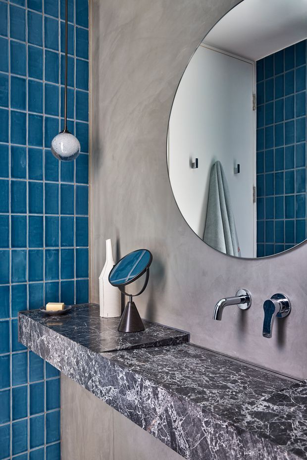 Close-up of a refined bathroom vanity featuring a dark veined stone benchtop, vivid blue square wall tiles, a grey plastered wall, wall-mounted chrome tapware, and a large rounded mirror. Decorative accessories sit on the floating vanity, with a towel reflected in the mirror.
