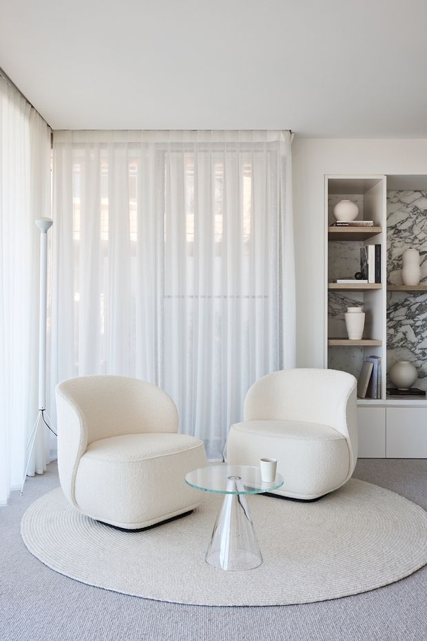 A bright, serene living room with floor-to-ceiling sheer curtains, two rounded cream boucle lounge chairs, a small glass coffee table, and a circular textured rug. A built-in shelving unit with dark timber shelves and a dramatic marble-look backing provides the main feature wall. The space has a soft, monochromatic neutral palette and clean contemporary detailing.