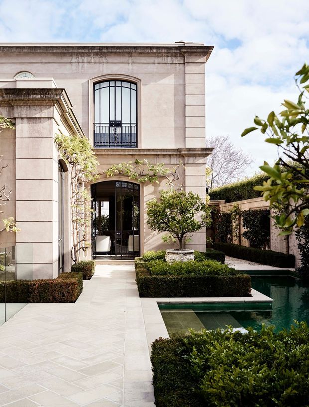 A formal landscaped courtyard beside a refined pale stone or rendered masonry building. The facade features tall black-framed arched glazing, classical horizontal cornices and pilasters, with French-style doors opening onto a light paved path. Carefully clipped hedges, topiary, espaliered planting and a narrow geometric reflecting pool create a symmetrical, elegant outdoor setting.