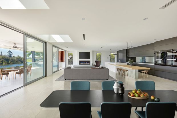 Bright open-plan contemporary living, dining and kitchen space with floor-to-ceiling glazing, skylights, a large dark dining table, upholstered dining chairs, a grey sofa, fireplace, island bench and extensive dark built-in cabinetry. A waterfront landscape is visible through the glazing, with a covered outdoor dining area beyond.