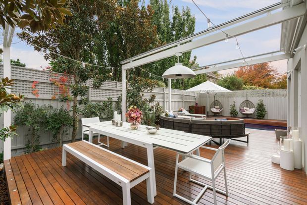 A landscaped outdoor entertaining deck enclosed by a white privacy screen and garden planting. The space features a white dining table with bench seating, an outdoor lounge with a low table, hanging egg chairs, timber decking, a covered pergola with exposed beams, and suspended lighting. The overall design is bright, relaxed, and well suited to outdoor dining and entertaining.