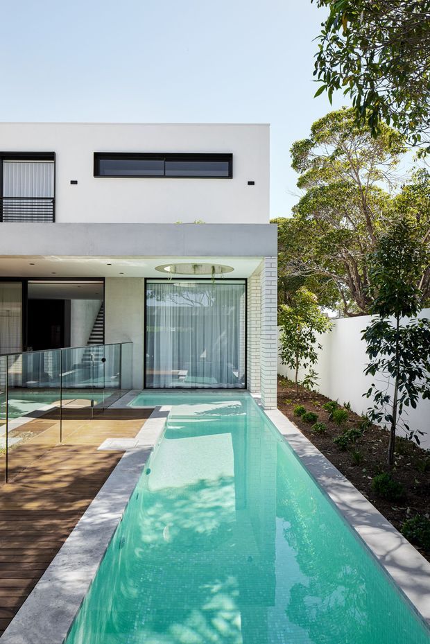 A contemporary multi-level residential home with a crisp white rendered facade, dark-framed windows, and extensive glazing opening onto a long narrow turquoise swimming pool. A timber deck borders the pool, while a landscaped garden and white boundary wall line the right side. The pool appears to extend beneath a covered outdoor area, with a circular ceiling opening adding a distinctive architectural feature.