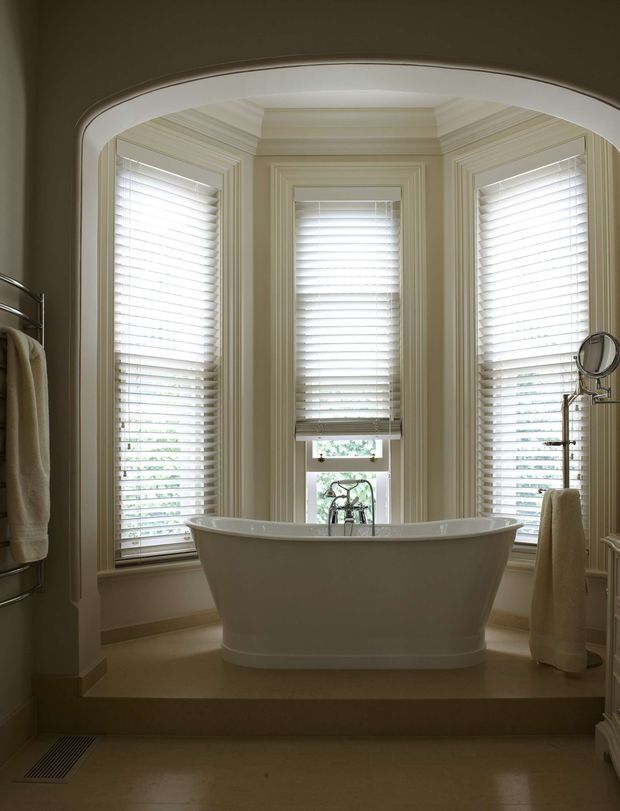 A refined bathroom featuring a freestanding white bathtub set on a raised platform within a curved bay window. Three tall windows have white plantation shutters and are surrounded by ornate layered trim and wall panelling. Neutral beige walls and flooring create a warm, traditional atmosphere, with towel rails and a small magnifying mirror visible at the sides.