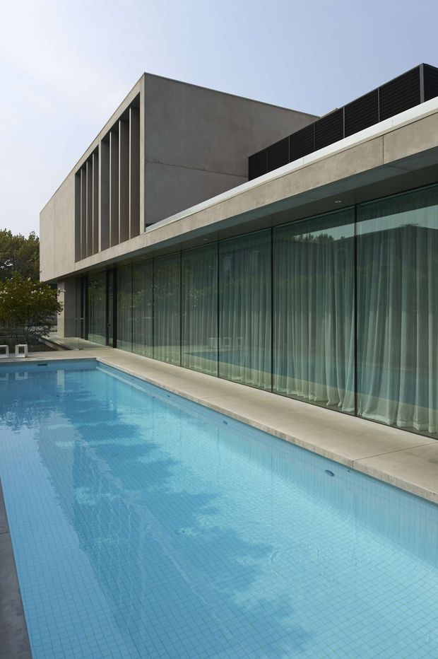 Exterior view of a minimalist modern two-storey residence beside a long rectangular swimming pool. The building has a smooth concrete facade, strong horizontal and vertical geometry, a projecting flat roofline, dark louvred screening, and extensive floor-to-ceiling glazed walls with sheer curtains. A pale paved pool surround and mature planting frame the scene.