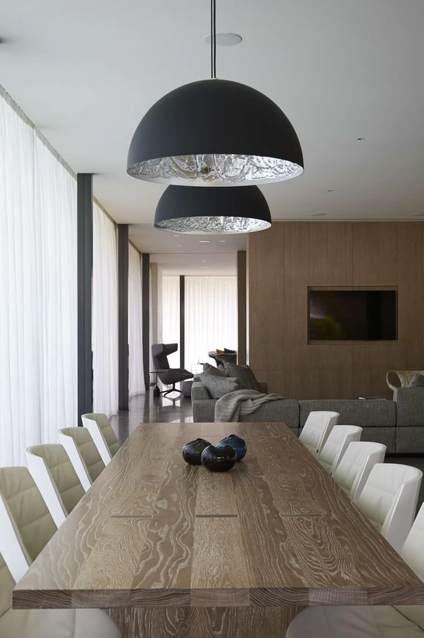 A refined open-plan dining and living space with a large rectangular natural timber dining table surrounded by white upholstered dining chairs. Two oversized black dome pendant lights with textured metallic interiors hang above the table. Behind it is a grey sectional sofa, contemporary lounge chairs, and a wall-mounted television set into a full-height timber feature wall. Floor-to-ceiling sheer curtains provide soft daylight, while dark slender window frames and recessed ceiling lighting reinforce the modern, minimalist character.