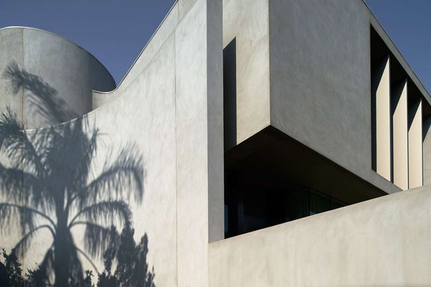 Exterior architectural photograph of a sculptural modern building with smooth pale rendered or concrete walls, strong geometric projections, deep recessed openings, and a curved cylindrical wall at the left. Palm shadows add contrast across the facade, while the dark blue sky frames the upper structure.