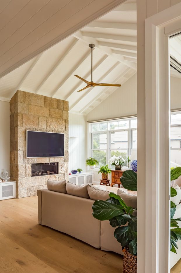 Bright, warm living room viewed through a doorway, featuring a dramatic white pitched ceiling with exposed beams and white wall panelling, a large natural stone fireplace with mounted television, pale timber flooring, a neutral sofa, abundant indoor plants, and generous white-framed windows. A timber ceiling fan and soft natural light contribute to the relaxed coastal character.