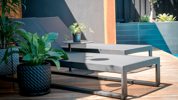 A sunlit contemporary outdoor deck or terrace furnished with two low-profile dark grey rectangular coffee tables, surrounded by lush potted plants. The setting features timber decking, dark rendered walls, a warm timber-framed opening, and blue-grey tiled or clad steps in the background. The furniture has clean, minimalist lines and a durable metal-like finish.