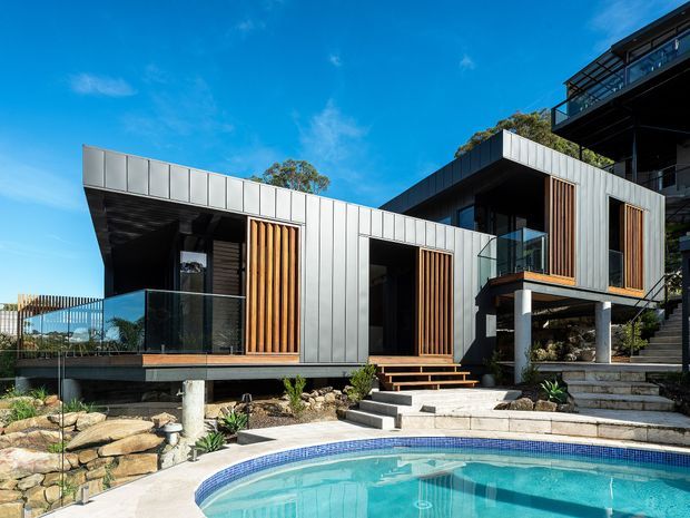 Contemporary multi-level residence with a strong geometric form, clad in light grey metal panels and accented with vertical timber screens. The house features projecting volumes, extensive dark-framed glazing, glass balustrades, raised decks, landscaped stone retaining walls, and a curved swimming pool in the foreground.