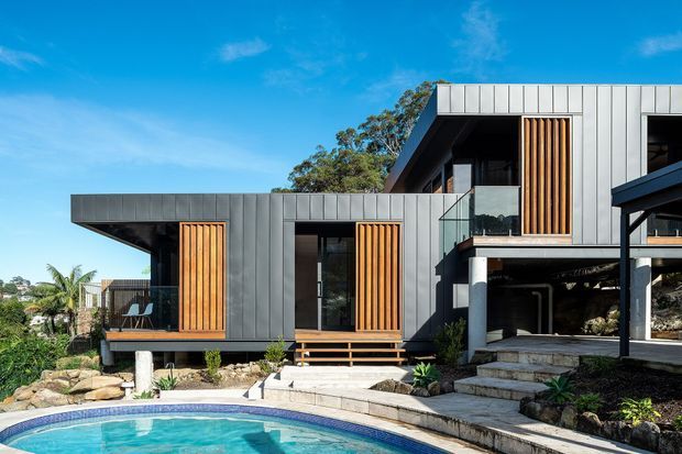 Exterior view of a contemporary multi-level residence with dark grey vertical metal cladding, projecting flat roof forms, extensive glazing, and warm timber privacy screens. The house is set on a landscaped sloping site with exposed structural supports, steps, rockwork, and a curved swimming pool in the foreground.