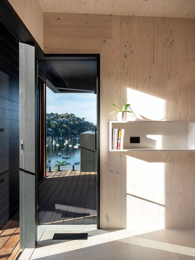 A bright, compact modern interior lined in pale timber, with a built-in white display shelf and black-framed doorway opening onto a timber deck. Through the doorway, a marina and waterfront hillside are visible. The space features strong sunlight, dark contrasting wall and door elements, and clean minimalist detailing.