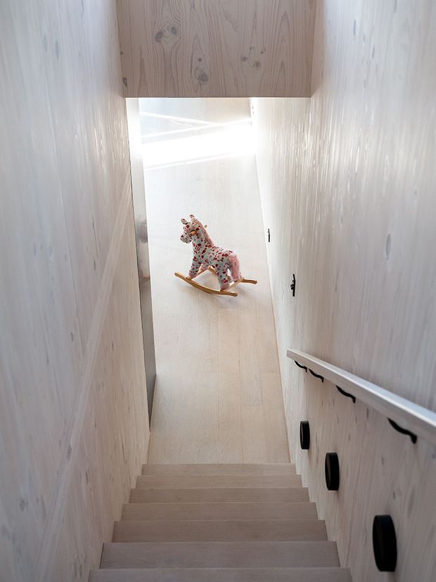 A narrow, light-filled timber-lined stairway with pale wood walls, stair treads, and a matching overhead beam. Black hardware and a simple handrail run along the right wall, leading to a small landing where a colorful rocking horse is displayed. The space has a clean, minimalist character with strong natural light.