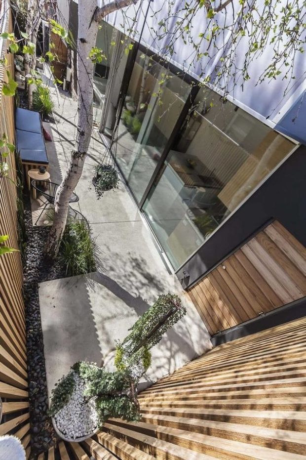 A modern residential courtyard viewed from above, with a narrow concrete-paved outdoor space framed by extensive floor-to-ceiling glazing, dark metal frames, timber decking and a broad timber-clad stair or stepped terrace. Mature trees, pebble garden beds, potted greenery and a small outdoor seating area create a leafy, private courtyard atmosphere.
