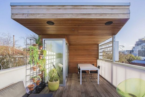 A compact rooftop or upper-level outdoor terrace with a timber-lined covered canopy, recessed downlights, timber decking, white privacy walls, and horizontal slatted screening. A glazed door opens into the interior, while potted plants, a small dining table, and chairs create a sheltered outdoor living and dining area. Urban buildings and trees are visible beyond the terrace.