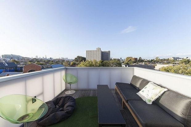 A bright rooftop terrace overlooking a dense urban neighbourhood. The terrace is enclosed by white privacy screens and furnished with a long dark outdoor sectional sofa, a narrow rectangular coffee table, a translucent green sculptural chair, and artificial turf flooring. The open sky and city skyline create an airy elevated setting.