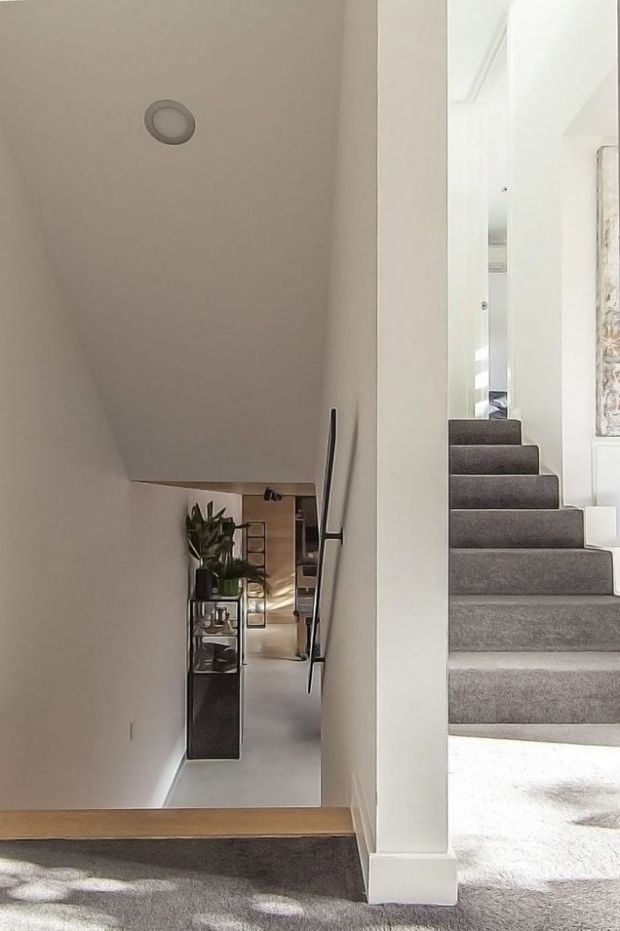 A bright, minimalist interior stairway and landing viewed from an upper level. Grey carpeted stairs descend through a narrow stairwell, with white painted walls, white trim, a simple dark handrail, and a timber stair nosing or landing edge. A hallway and console shelving with a plant are visible below, while another flight of carpeted stairs rises at the right.