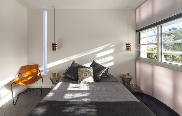 Bright contemporary bedroom with a neatly made bed, dark grey bedding and layered cushions against a plain light wall. Two slim pendant lights hang over mismatched bedside tables, with a sculptural orange lounge chair near the window. Strong sunlight and linear shadows enter through large horizontal windows fitted with roller blinds, creating a clean, relaxed atmosphere.