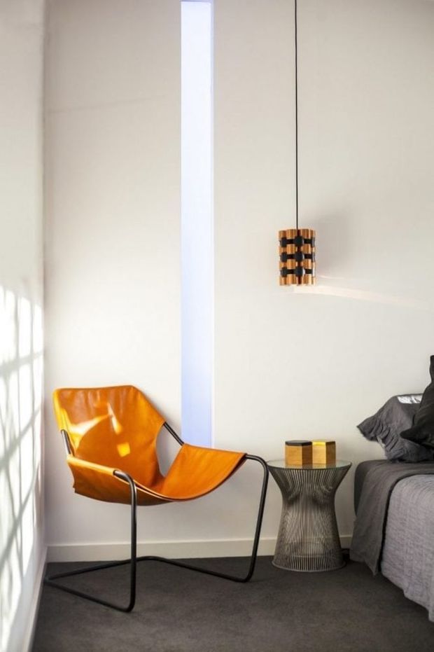 A bright, minimalist bedroom corner with white walls, a narrow vertical window, dark grey carpet, and a black upholstered bed partially visible at right. A vivid orange leather sling chair with a black metal frame sits beside a sculptural wire-base side table, while a small cylindrical pendant light with warm wood and dark bands hangs above.