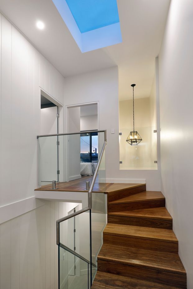 A bright modern stairway with richly grained dark timber treads and landing, framed by clear glass balustrades with slim metal handrails. White vertical wall panelling and plaster walls create a clean backdrop. A large blue-tinted skylight brings daylight from above, while a geometric pendant light hangs in a double-height void beside the stairs. An open doorway reveals part of an adjacent bedroom or room.