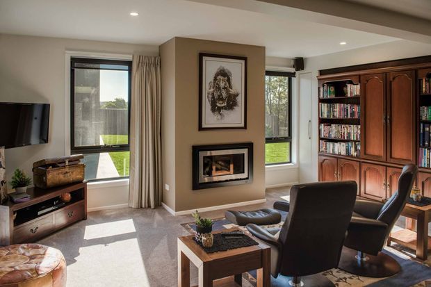 A warm, well-appointed living room with large windows, neutral walls, carpeted flooring, a central fireplace beneath framed artwork, a wall-mounted television, and a substantial dark timber built-in bookcase. Two dark leather lounge chairs and a small timber coffee table create a comfortable reading and seating area.