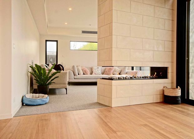 Warm contemporary open-plan living area featuring a large tiled fireplace with a long horizontal firebox, pale upholstered seating, a potted plant, carpeted lounge zone, and light timber flooring in the foreground. Black-framed glazing and simple recessed ceiling lighting complete the clean, refined interior.