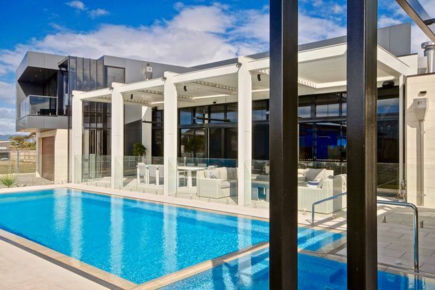A bright contemporary house exterior centered on a rectangular swimming pool and poolside entertaining area. White rendered walls, dark metal-framed floor-to-ceiling glazing, deep outdoor pergola structures, and a projecting upper-level volume create a modern architectural composition. The covered terrace contains pale outdoor seating and dining furniture, with glass pool fencing and light paving around the water. A second building wing is visible beyond the terrace.