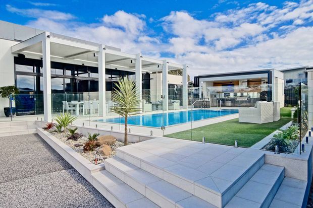 A contemporary outdoor entertainment area with a rectangular swimming pool, extensive frameless glass balustrades, pale tiled terraces and broad steps. White structural columns support covered open-air spaces, with a modern pavilion and outdoor lounge seating beyond the pool. Gravel landscaping, raised planters and sculptural drought-tolerant plants surround the bright, open courtyard.