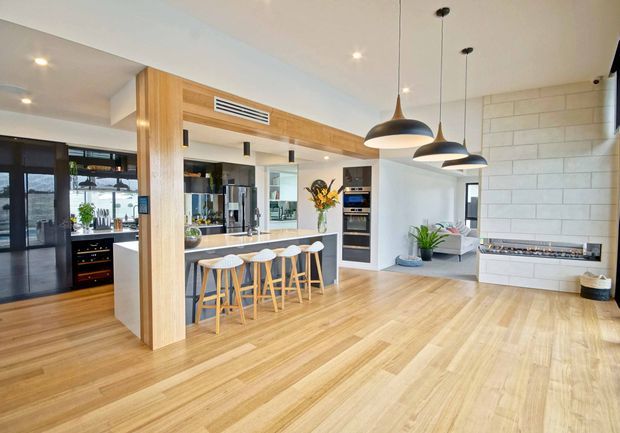 Open-plan contemporary kitchen and living area with a large timber island and integrated cabinetry, five bar stools, stainless-steel appliances, pale timber flooring, white tiled walls, and three black pendant lights. A built-in fireplace or media unit is visible on the right, with glazing and dining/living areas beyond the kitchen.