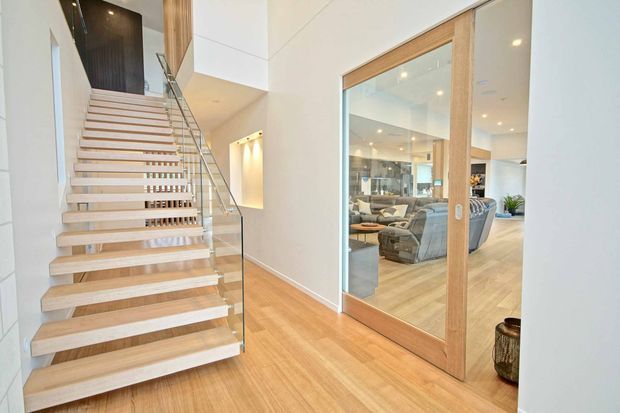 Bright contemporary interior view of a hallway and open stairway. The space features pale timber flooring, a floating timber staircase with a clear glass balustrade, white walls, recessed lighting, and a large timber-framed glazed sliding door revealing a living room beyond.