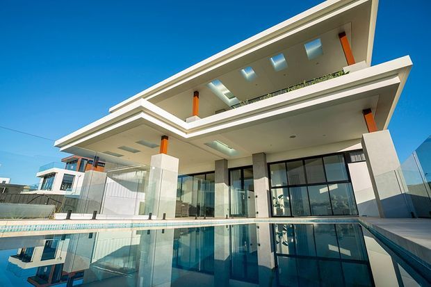 Modern multi-level residential exterior viewed from beside a reflective swimming pool. The building features crisp white cantilevered slab forms, extensive floor-to-ceiling glazing, exposed orange structural columns, roof skylights, glass balustrades, and a bright blue-sky setting.