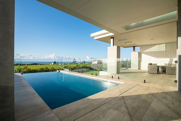 A covered modern outdoor entertaining area opens onto a rectangular swimming pool, with large tiled flooring, square columns, recessed ceiling lighting, and a skylight. Beyond the landscaped grounds is a broad coastal or lakeside view with distant snow-capped mountains.